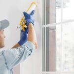A man in a cap and gloves applying caulk beside a white-framed window in a bright room with large glass panes.