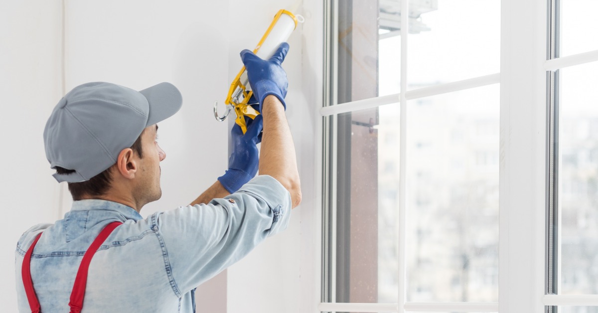 A man in a cap and gloves applying caulk beside a white-framed window in a bright room with large glass panes.