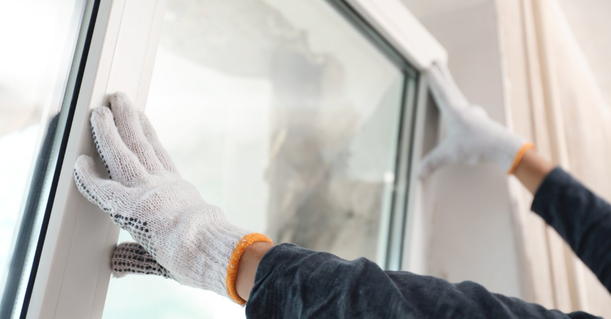 Gloved hands pressing a white window frame beside glass panes and light curtains during indoor installation.