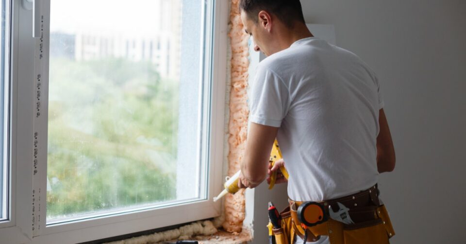 A man using a caulking gun around a white window frame with exposed foam, tools, and a yellow tool belt.