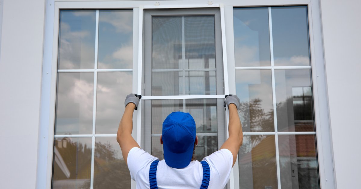 A person in a blue cap and gray gloves lifting a white framed screen onto a large window outside a building.