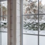 A pair of white interior windows looking toward snow-covered shrubs and trees outside a wood-paneled room.