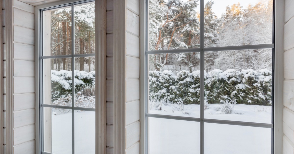 A pair of white interior windows looking toward snow-covered shrubs and trees outside a wood-paneled room.