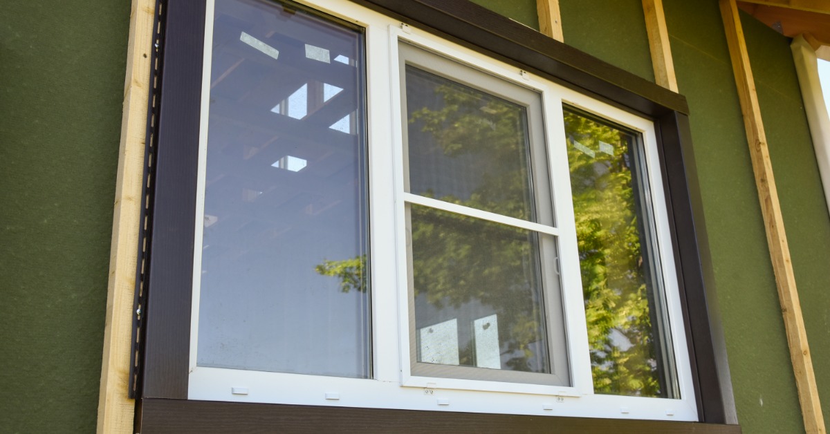 A white window installed in a wall with green sheathing, exposed wood studs, and dark exterior trim.
