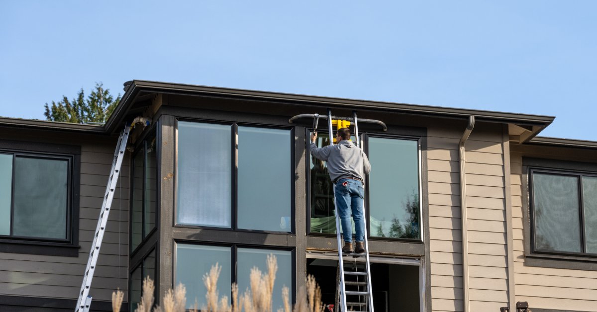 A worker standing on an extension ladder beside a large two-story house with windows, siding, and a blue sky.