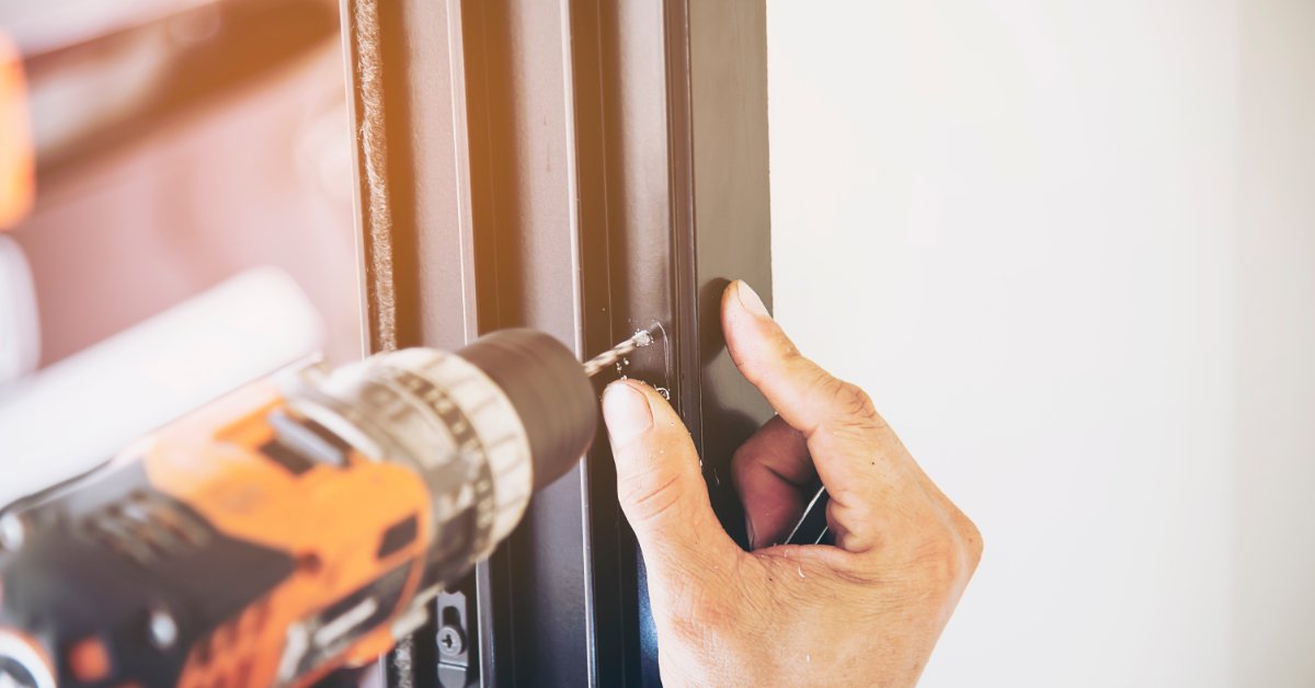 A worker installing an aluminum screen frame with a screwdriver beside glass panels at a construction site.