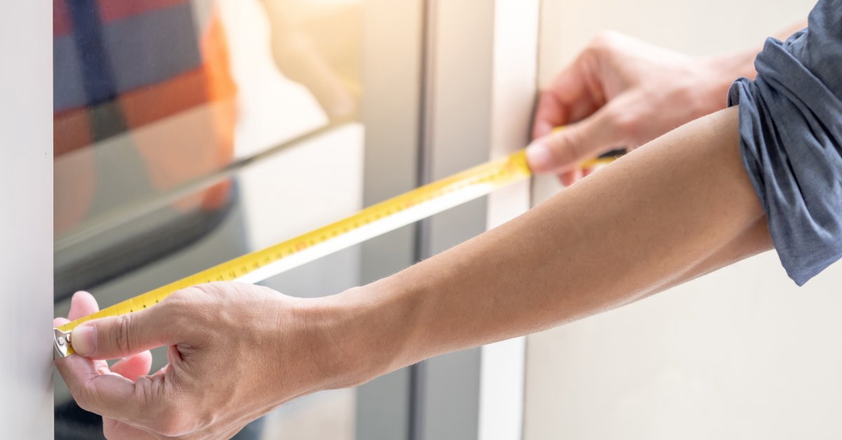 Hands using a yellow tape measure along an aluminum window frame with tools and materials on a work surface.