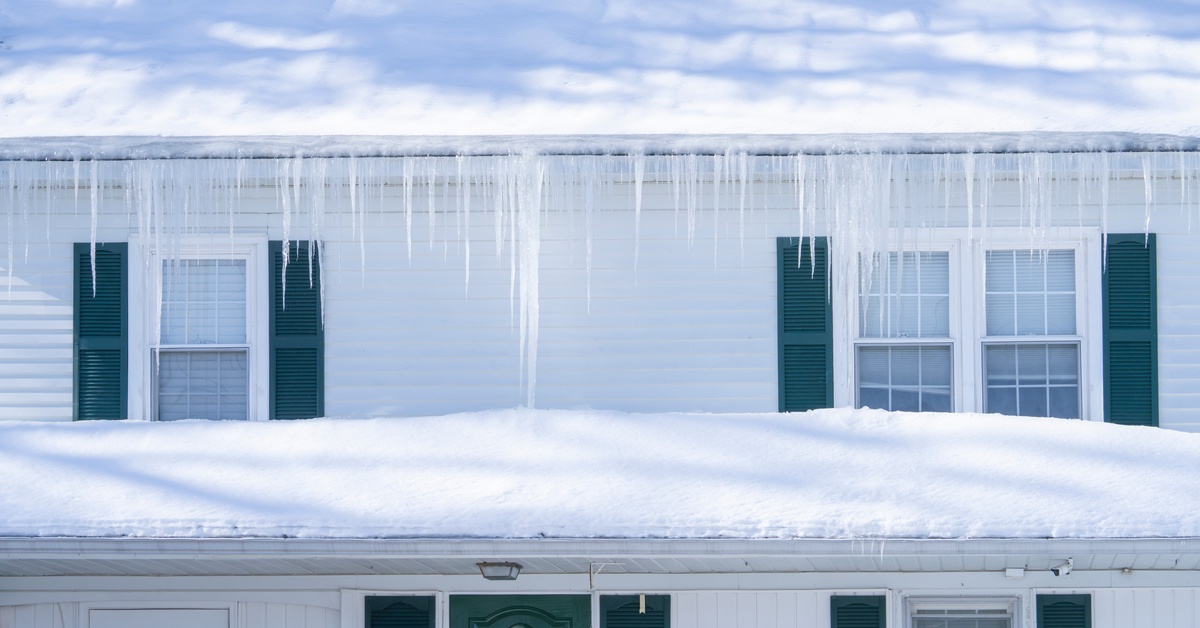 A snowy house roof with icicles hanging along the edge near exterior windows and siding on a winter day.
