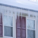 A house roof covered with snow and long icicles hanging from the eaves above exterior windows in winter.