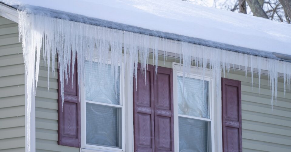 A house roof covered with snow and long icicles hanging from the eaves above exterior windows in winter.