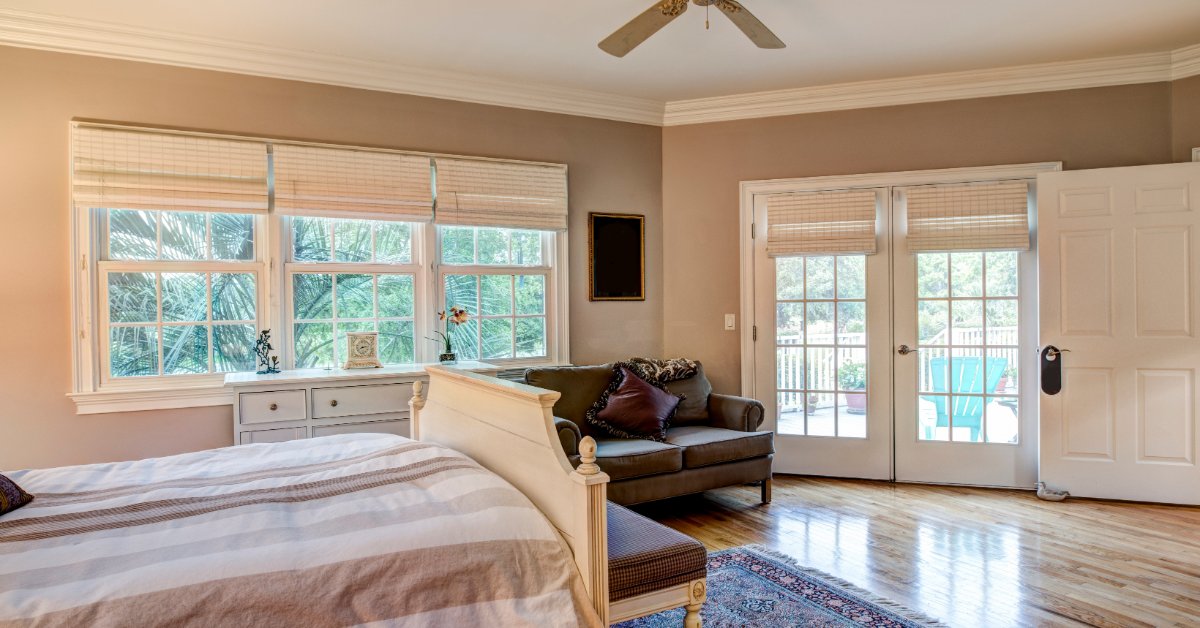 A bedroom with hardwood floors, French doors, a white bed, gray bench, patterned rug, and beige curtains.