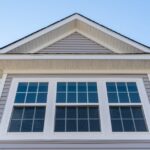 A white double-hung window with grille panes set in horizontal grey vinyl siding beneath a gable and soffit.