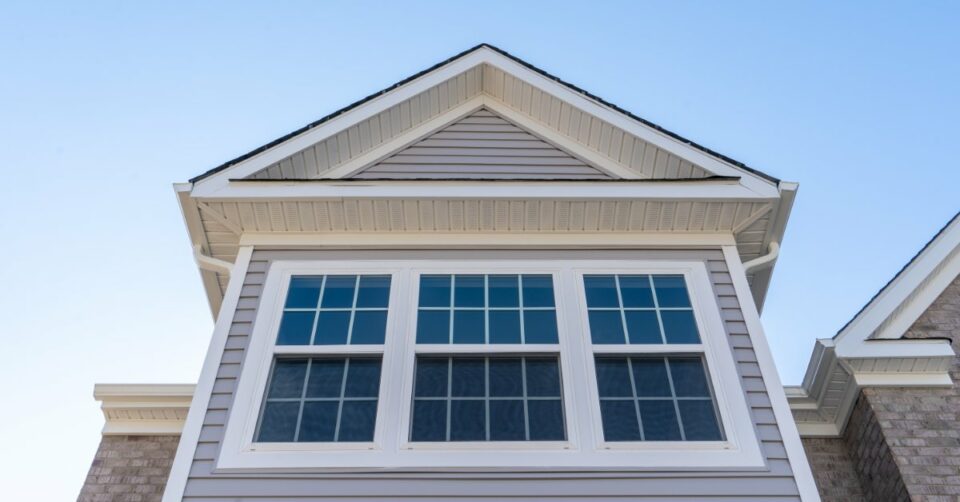 A white double-hung window with grille panes set in horizontal grey vinyl siding beneath a gable and soffit.