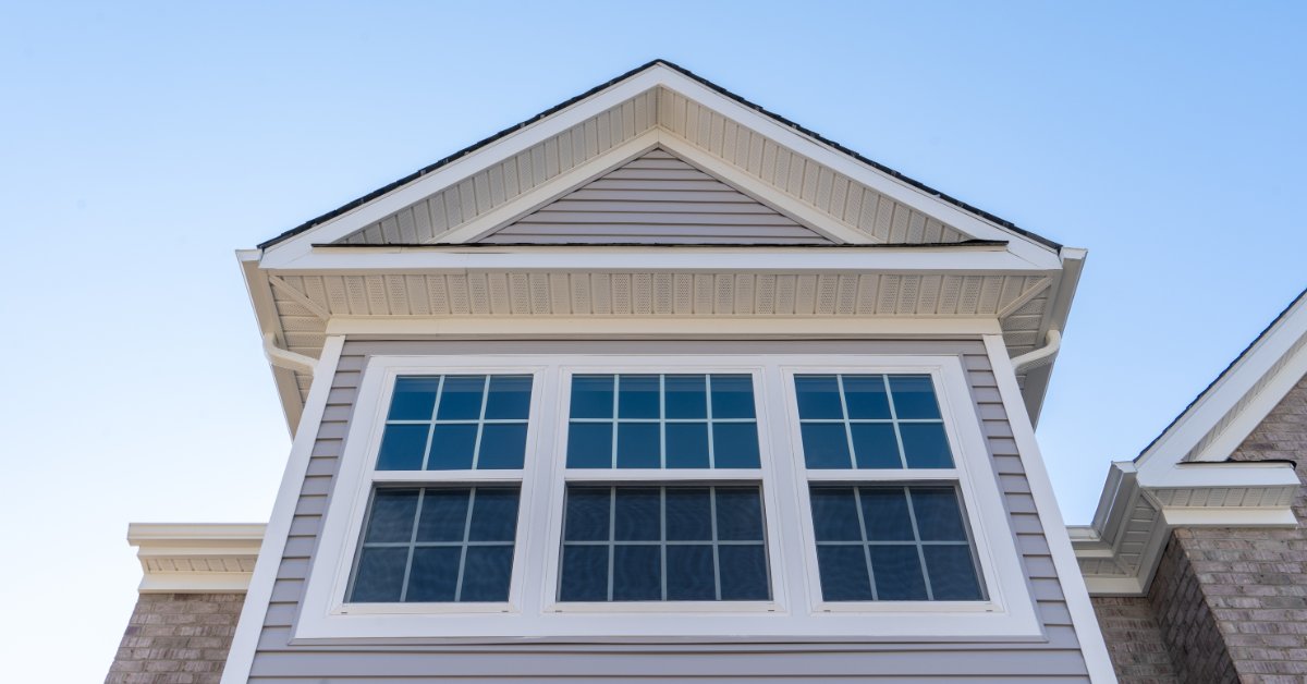 A white double-hung window with grille panes set in horizontal grey vinyl siding beneath a gable and soffit.