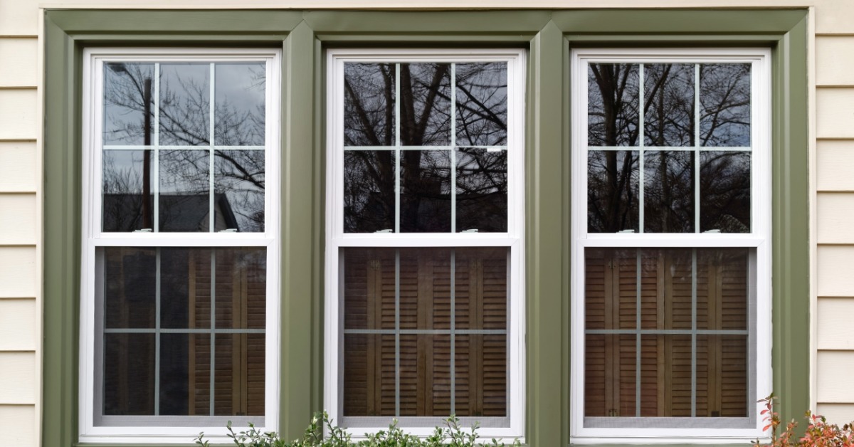 White replacement windows with green trim set into horizontal siding, with reflections visible in the glass.