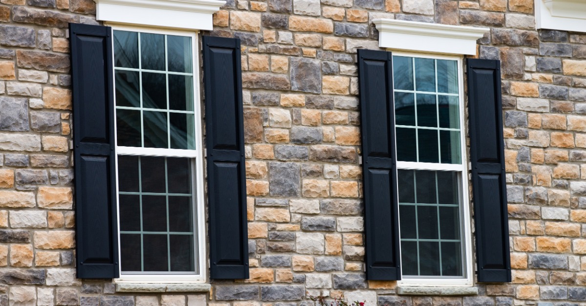 A two-pane white window with dark shutters set in light vinyl siding, with roof shingles along the top.