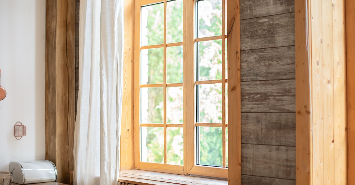 A tall wooden window with divided panes, a white curtain, and stacked firewood beneath its sill beside a timber wall.
